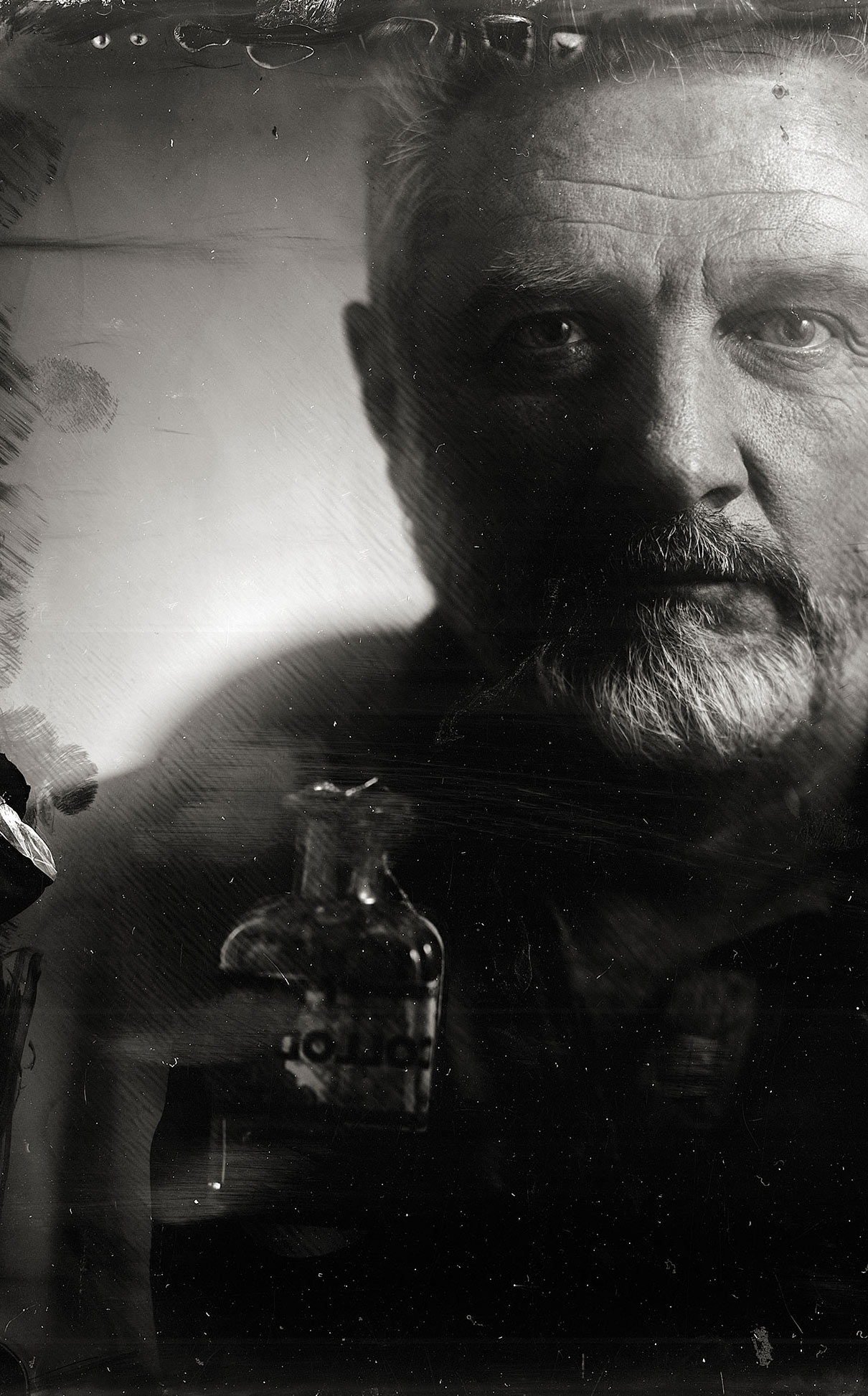 A black and white portrait of an older man with a serious expression, partially obscured by a glass bottle in the foreground. The image has a vintage, textured appearance reminiscent of the tintype old photography technique.