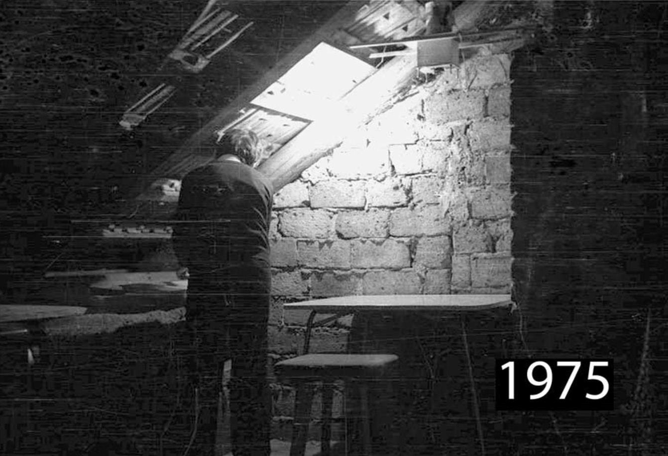 A person stands under a slanted roof near a brick wall, illuminated by light from a window above. The room is dimly lit and rustic. The year 1975 appears in bold in the bottom right corner. It is improvised darkroom.