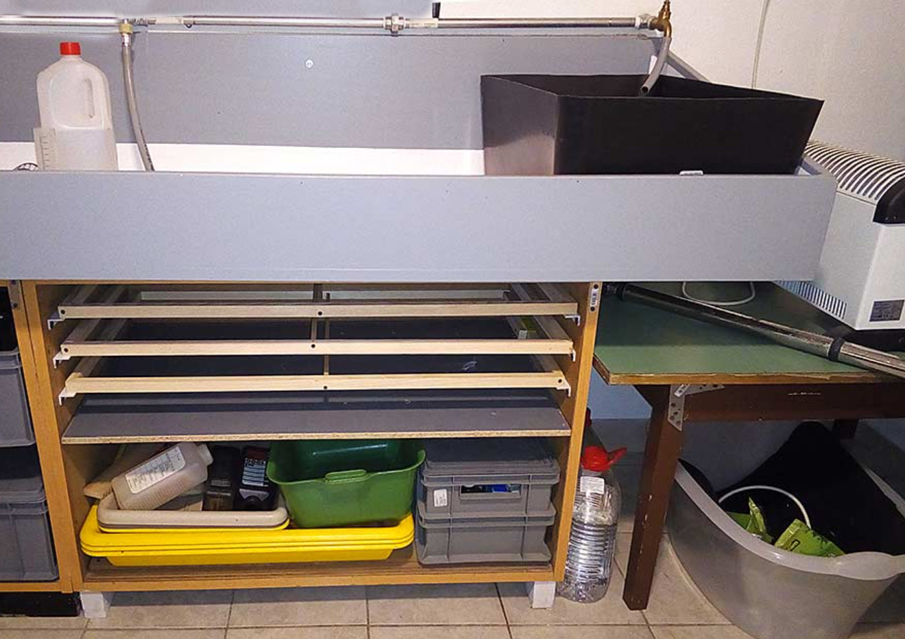 A utility sink setup with a black basin, shelves holding trays, bins, and containers, and various cleaning supplies underneath. A green table with equipment is beside the sink on a tiled floor.