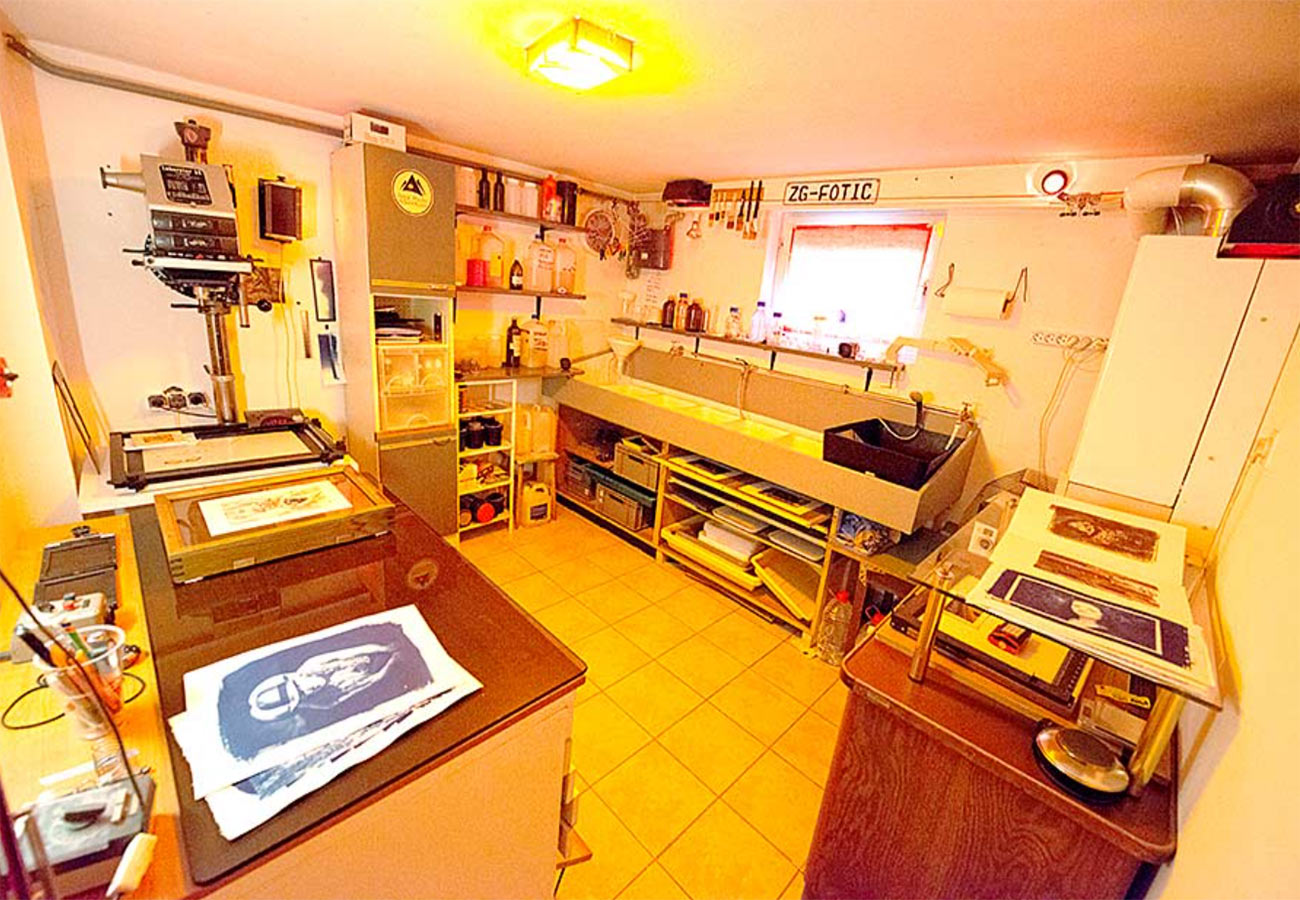A brightly lit photographic darkroom with yellow-tiled floor, print dryer, enlarger, various trays, bottles, chemical containers, and prints drying on countertops and racks. A small window lets in some natural light.